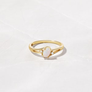 May include: A gold ring featuring an oval opal center stone and two small accent diamonds. The ring has a split shank design. The opal has a milky white color, and the diamonds sparkle. The ring is set against a white marble background.