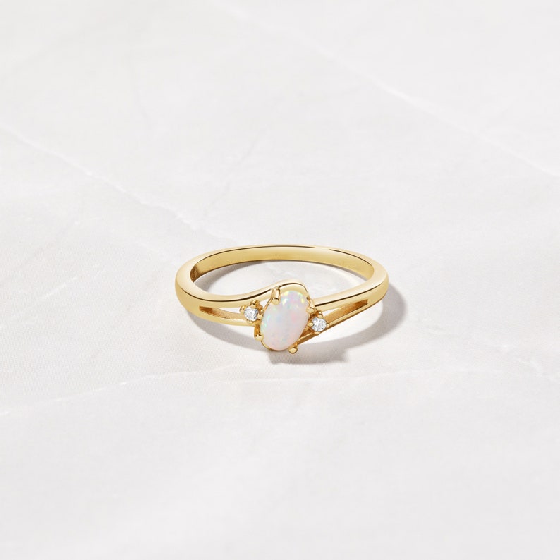 May include: A gold ring featuring an oval opal center stone and two small accent diamonds. The ring has a split shank design. The opal has a milky white color, and the diamonds sparkle. The ring is set against a white marble background.