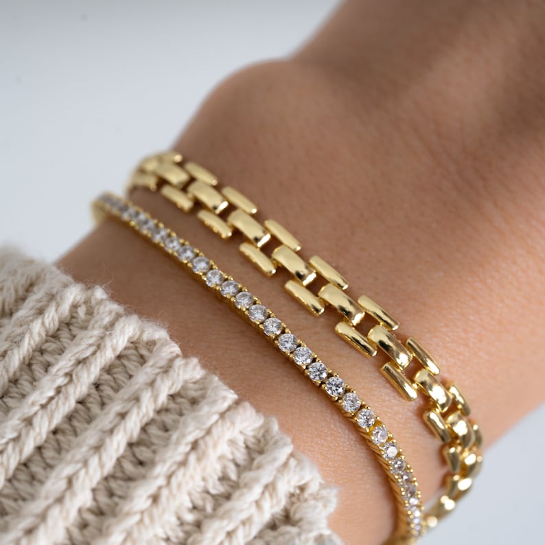 Pode incluir: Duas pulseiras de ouro em um pulso. Uma pulseira &eacute; uma corrente delicada com pequenas pedras transparentes. A outra pulseira &eacute; uma corrente mais grossa com um design mais intrincado.