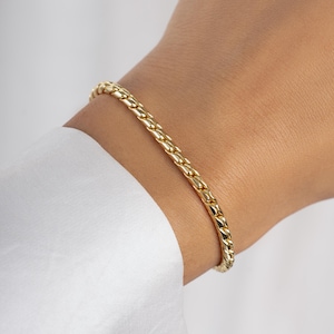 May include: A gold-colored curb chain bracelet. The bracelet is made of interlocking links and is worn on a wrist. The wrist is covered by a white long-sleeved shirt.