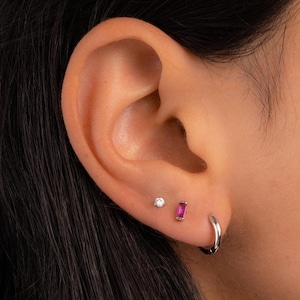 Może przedstawiać: Zbliżenie ucha z trzema kolczykami: małym diamentem, prostokątnym rubinem i srebrną kolczykiem kołem. Ucho na ciemnym tle.