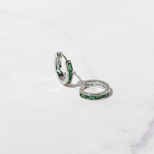 May include: A pair of silver hoop earrings with a row of emerald green baguette-shaped stones around the edge.