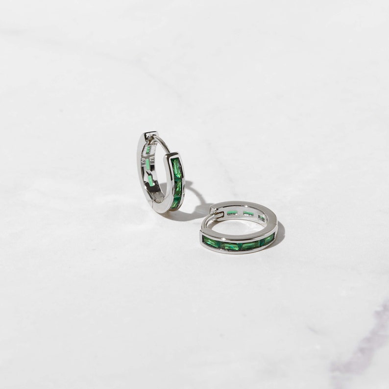 May include: A pair of silver hoop earrings with a row of emerald green baguette-shaped stones around the edge.