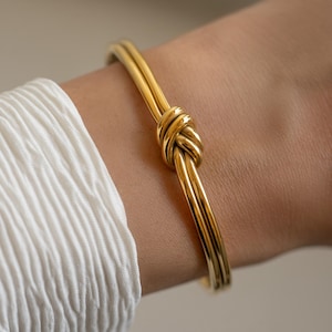 Puede incluir: Una pulsera rígida de tono dorado pulido con un diseño de nudo central. La pulsera está construida con dos bandas metálicas lisas y redondeadas que convergen en un único nudo decorativo en el centro, creando una superficie continua, elegante y brillante.