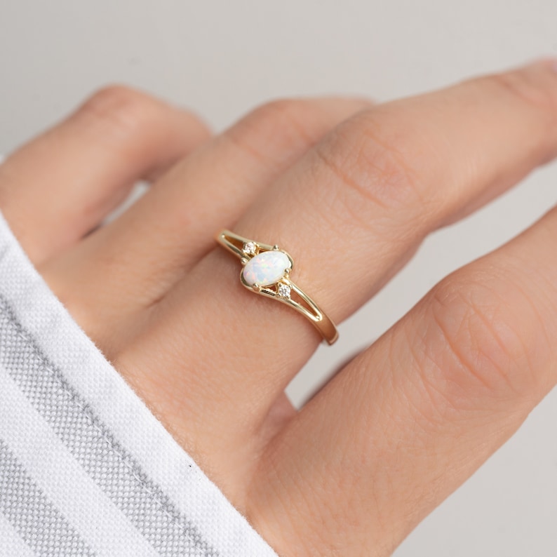 May include: A gold ring featuring an oval opal gemstone and two smaller accent stones. The ring has a delicate, open design with a polished finish. The opal has a milky white appearance. The ring is a piece of jewelry.