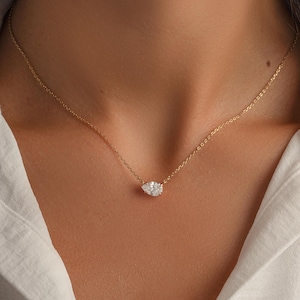 May include: A delicate gold necklace featuring a pear-shaped, clear gemstone pendant. The pendant is centered on a thin gold chain, resting gracefully against a white shirt. The necklace is a simple yet elegant piece of jewelry.
