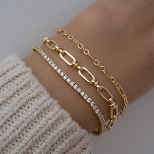 Pode incluir: Tr&ecirc;s pulseiras de ouro empilhadas em um pulso. A pulseira superior &eacute; uma corrente delicada, a pulseira do meio &eacute; uma corrente grossa com elos ovais, e a pulseira inferior &eacute; uma corrente fina com pequenas pedras transparentes.
