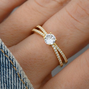 May include: A gold ring with a single round clear stone in the center. The ring has two bands that cross over each other and are set with small clear stones.