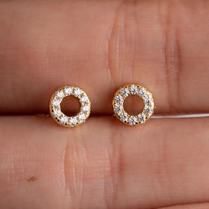 May include: A pair of gold-colored stud earrings. Each earring features a circular design encrusted with small, clear gemstones. The earrings are set against a skin-toned background.