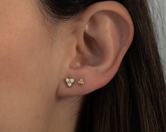 May include: A pair of gold earrings featuring small white opal stones. One earring is in the shape of a triangle, the other is a cluster of three small balls.