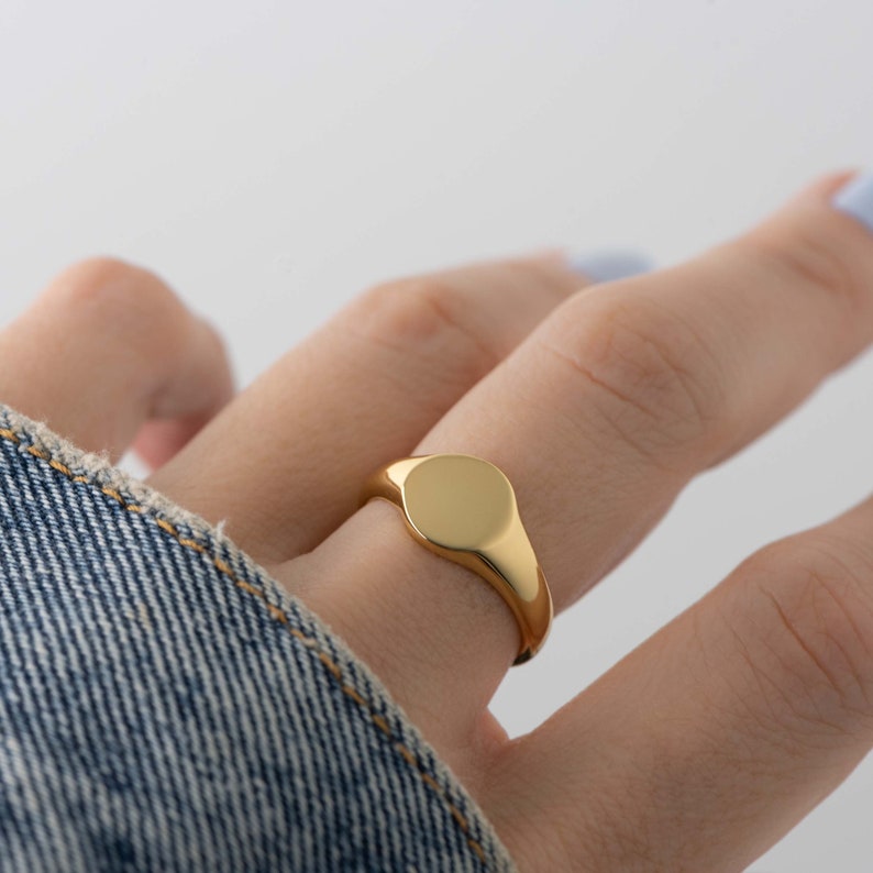 May include: A gold signet ring with a smooth, circular face. The ring is worn on a finger, with a denim jacket sleeve visible in the background. The ring's polished surface reflects light, highlighting its simple, elegant design.