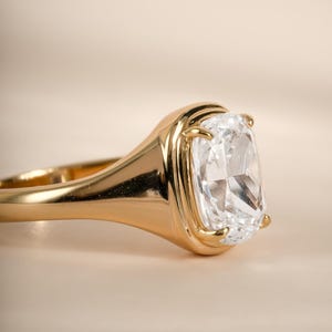 Elongated diamond ring in gold with a cushion cut stone set in a bezel and wide tapered band.