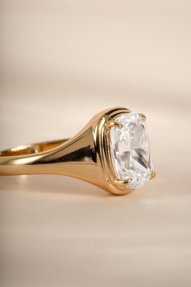 Elongated diamond ring in gold with a cushion cut stone set in a bezel and wide tapered band.