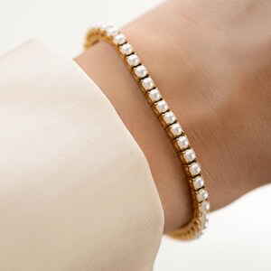 May include: A gold-toned tennis bracelet featuring a continuous row of small, round, white pearls set in individual metal settings. The bracelet encircles the wrist, showcasing a uniform design with a polished finish and a classic, linked structure.