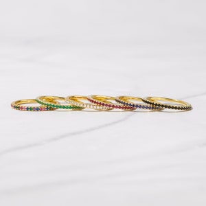 May include: Six gold rings with a single row of colorful gemstones. The rings are arranged in a row, with the black gemstone ring on the right and the green gemstone ring on the left.