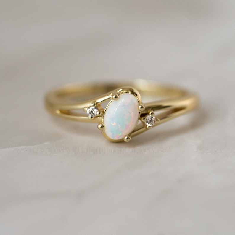 May include: A gold ring with an oval opal center stone and two small accent diamonds. The ring has a split shank design and is set on a white surface. The opal has a milky white color with flashes of blue and pink.