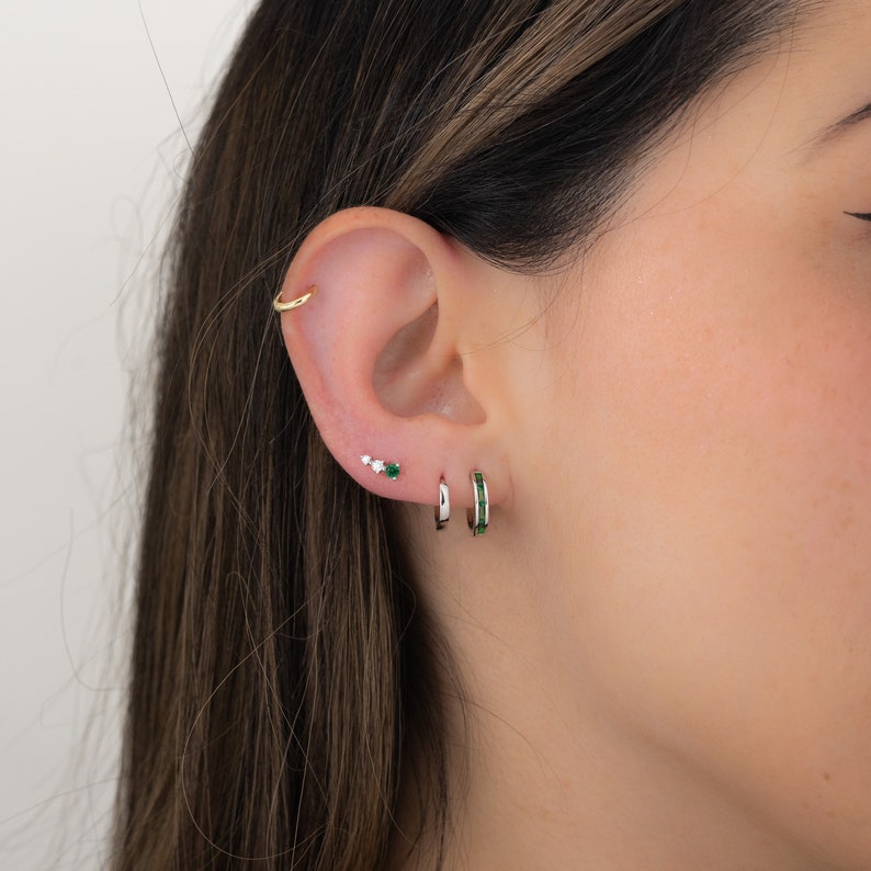 May include: A close-up of a person's ear with three earrings. The earrings are small and silver. The top earring is a cluster of three small diamonds. The middle earring is a single green gemstone. The bottom earring is a silver hoop with a row of green gemstones.