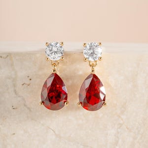 May include: A pair of gold-tone drop earrings featuring a round, clear, faceted stone at the top and a larger, pear-shaped, deep red faceted stone suspended below. The stones are held in place by gold-tone metal prongs, creating a classic dangle design.