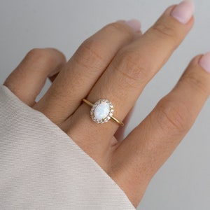 Pode incluir: Um anel de ouro com uma opala oval branca cercada por um halo de pedras transparentes menores.
