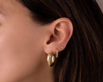 May include: A close-up of a person's ear adorned with three gold earrings. The set includes a pair of thick, polished hoop earrings, a smaller beaded hoop, and a tiny stud earring. The earrings are a bright, shiny gold color.