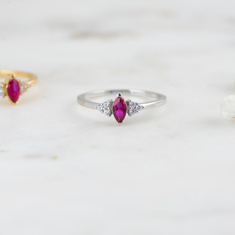 K&ouml;nnte beinhalten: Ein silberner Ring mit einem rubinroten Marquise-Schliff und kleinen, klaren Edelsteinen auf der Ringschiene.