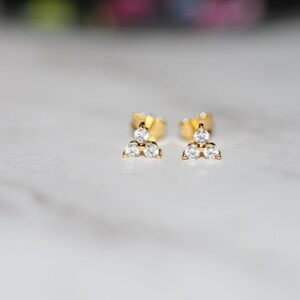 May include: A pair of gold stud earrings with three small, clear cubic zirconia stones arranged in a triangle shape.