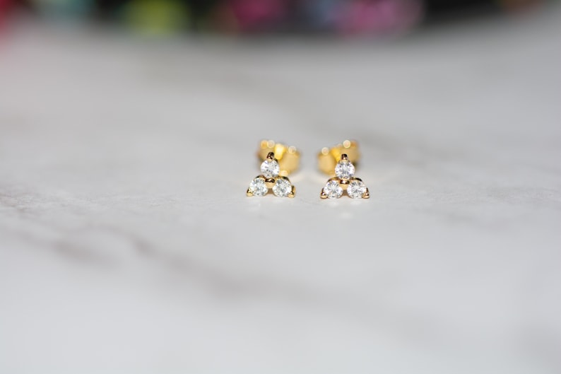 May include: A pair of gold stud earrings with three small, clear cubic zirconia stones arranged in a triangle shape.