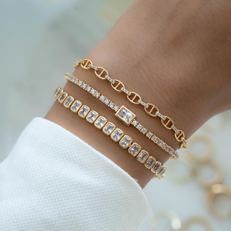 model wearing a chain cz gold bracelet, a tennis bracelet with a bezel emerald cut charm and a bezeled emerald cut tennis bracelet