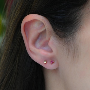 以下が含まれることがあります： 小さなイヤリングを2つつけた耳のクローズアップ。左のイヤリングは金のボール、右のイヤリングはピンクの宝石が3つ付いた小さな三角形です。