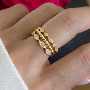 Peut inclure: Trois bagues dorées empilées sur un doigt. Les bagues présentent un motif torsadé et des pierres précieuses claires serties. Les bagues sont de couleur or chaud et se détachent sur un fond doux.