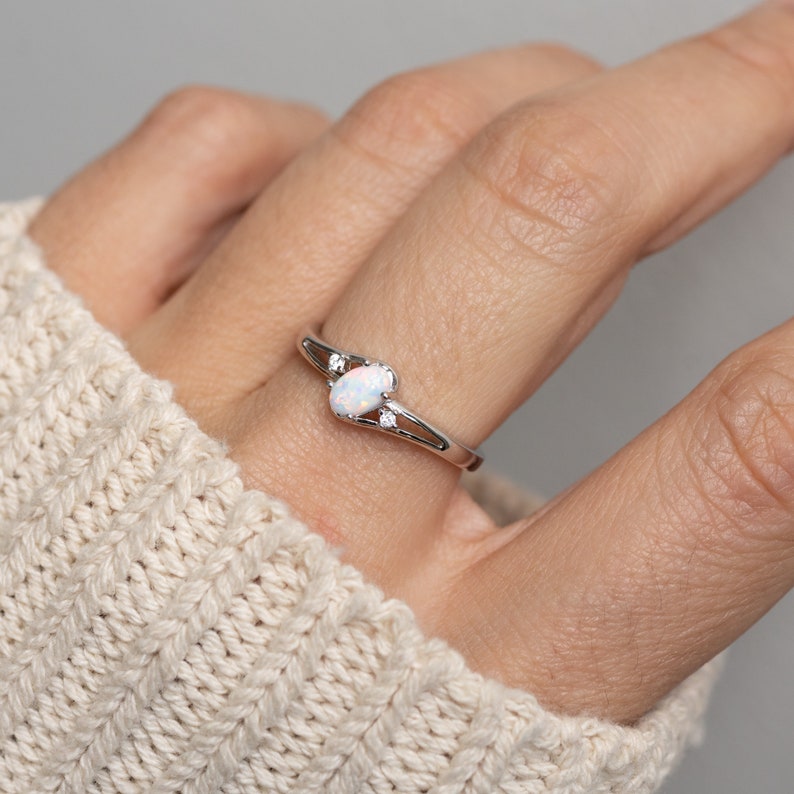 May include: A silver ring with an oval, white opal center stone and two small, round accent stones. The ring has a delicate, swirling design. The ring is shown on a person's finger, with a cream-colored sweater in the background.