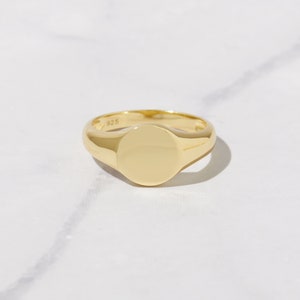 May include: A gold-colored signet ring with a polished, rounded face. The ring is made of a shiny, reflective metal and sits on a white surface. The ring is a classic piece of jewelry.