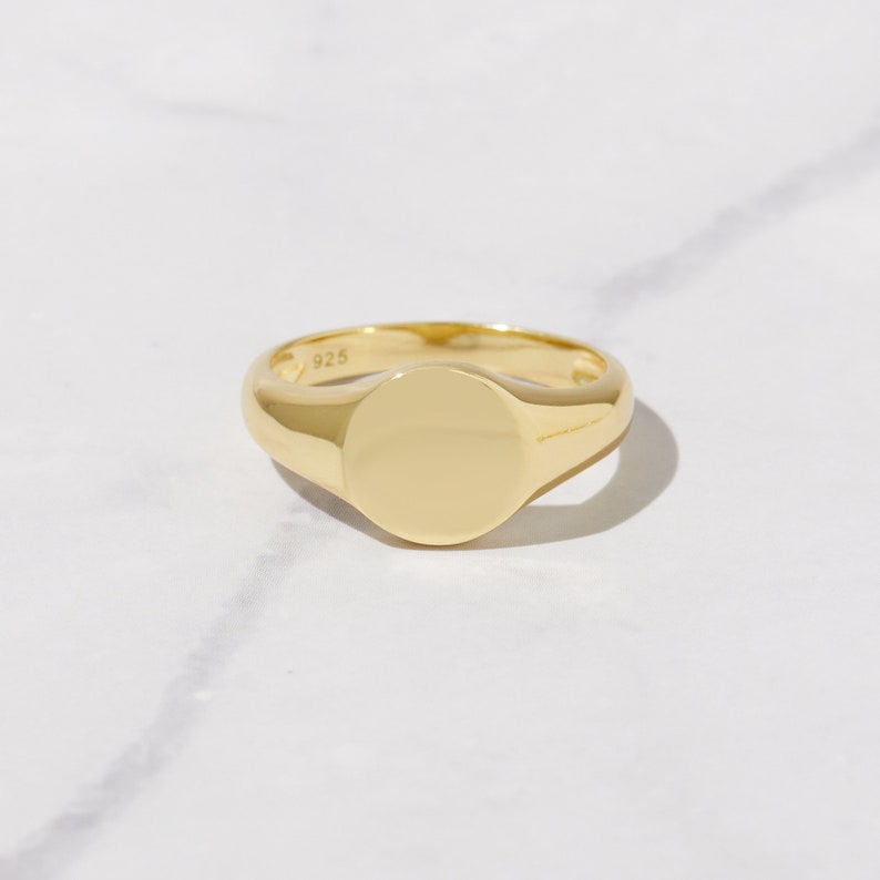 May include: A gold-colored signet ring with a polished, rounded face. The ring is made of a shiny, reflective metal and sits on a white surface. The ring is a classic piece of jewelry.
