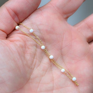 May include: A delicate gold chain necklace with small white opal beads. The necklace is layered with two chains and has a simple design.