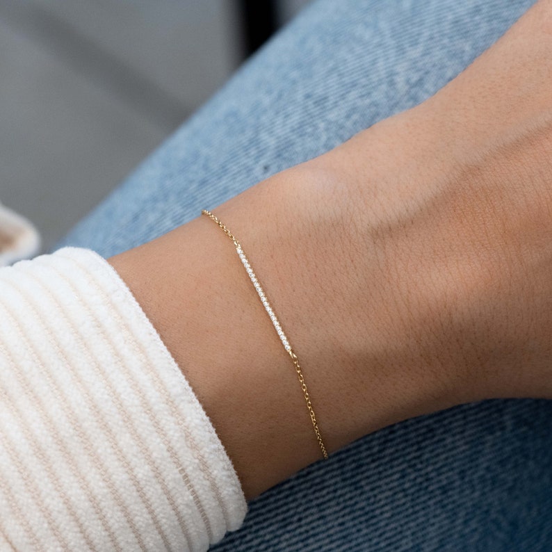 May include: A delicate gold chain bracelet with a thin bar of pave diamonds. The bracelet is worn on a person's wrist.