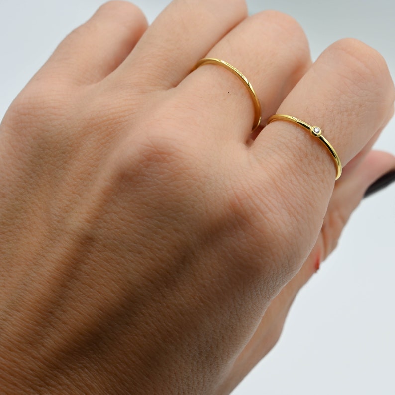 May include: Two gold rings on a hand. The ring on the index finger is plain, while the ring on the middle finger has a small, clear stone.