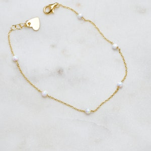 May include: A delicate gold chain bracelet with small white opal beads. The bracelet has a heart-shaped clasp.