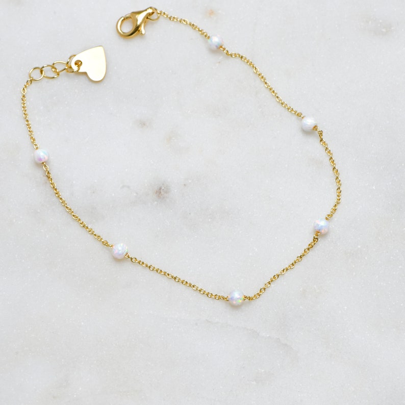 May include: A delicate gold chain bracelet with small white opal beads. The bracelet has a heart-shaped clasp.