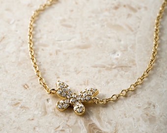 Op de afbeelding: Een goudkleurige ketting met een vlinderhanger. De vlinder is bezet met kleine, heldere edelstenen. De ketting is delicaat en heeft een schakelontwerp. De sieraden worden gepresenteerd op een lichtgekleurd, getextureerd oppervlak.