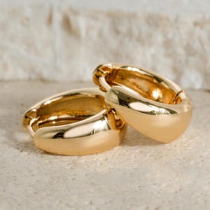 May include: Two gold-colored hoop earrings. The earrings are thick and have a smooth, polished surface. The earrings are resting on a light-colored, textured surface. The earrings are a classic jewelry item.