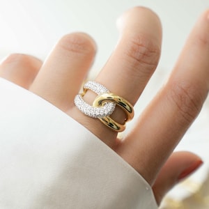 May include: A gold-toned ring with a chain link design. One link is encrusted with small, sparkling white stones, while the other is smooth gold. The ring is worn on a finger, showcasing its elegant design.