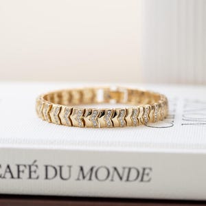 Puede incluir: Una pulsera dorada con diseño de chevrón, adornada con pequeñas piedras brillantes. La pulsera está sobre un libro con la inscripción "CAFÉ DU MONDE". La joya tiene una estética clásica y elegante.