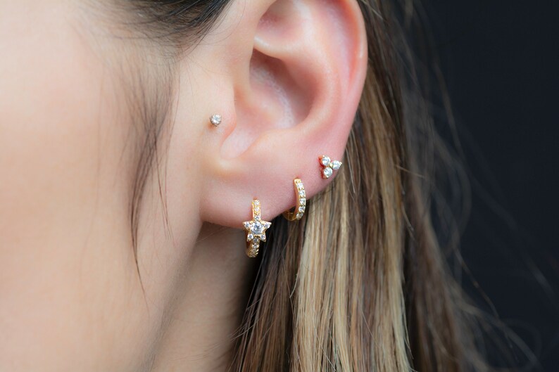 May include: A person's ear with multiple gold earrings. The earrings are set with small, clear stones. One earring is a star shape, another is a small hoop, and the third is a cluster of three stones.