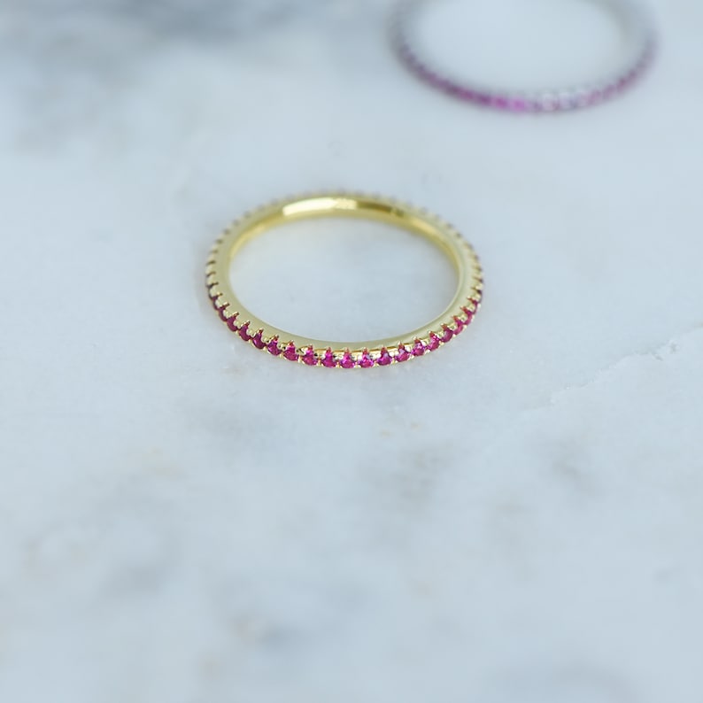 May include: A gold band ring with a row of small, bright pink gemstones around the entire band.