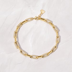 May include: A gold chain necklace with a heart-shaped clasp. The chain is made of small, square links that are set with clear cubic zirconia stones.