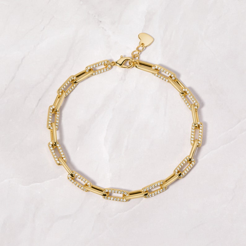 May include: A gold chain necklace with a heart-shaped clasp. The chain is made of small, square links that are set with clear cubic zirconia stones.