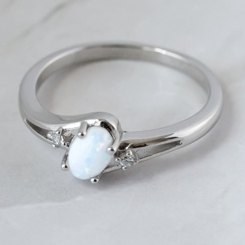 May include: A silver ring featuring an oval, white opal gemstone. The ring has a sleek, modern design with two small accent stones on either side of the opal. The band is smooth and polished.
