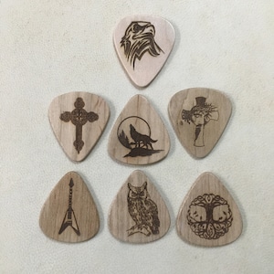 May include: A collection of seven wooden guitar picks, each with a unique engraved design. The designs include an eagle, a cross, a howling wolf, a cross with a face, an electric guitar, an owl, and a tree of life. The picks are light brown.