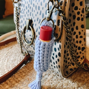 K&ouml;nnte beinhalten: Ein hellblau geh&auml;kelter Lippenbalsamhalter mit Quaste, befestigt an einer Handtasche mit Dalmatiner-Muster. Der Lippenbalsam hat eine rote Kappe. Die Handtasche hat einen braunen Riemen und goldfarbene Beschl&auml;ge.