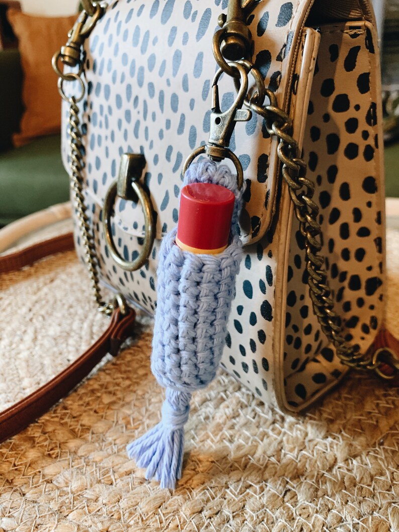 K&ouml;nnte beinhalten: Ein hellblau geh&auml;kelter Lippenbalsamhalter mit Quaste, befestigt an einer Handtasche mit Dalmatiner-Muster. Der Lippenbalsam hat eine rote Kappe. Die Handtasche hat einen braunen Riemen und goldfarbene Beschl&auml;ge.